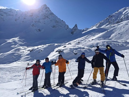 6 skieurs en tenus, équipés de skis ou de surf, se tiennent en rang face à l'objectif et lèvent les bras en signe de joie. Le paysage est couvert de neige et le soleil brille intensément derrière les montagnes.